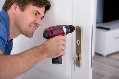 Close-up of a happy, smiling young man installing a lock on a door using a cordless screwdriver.